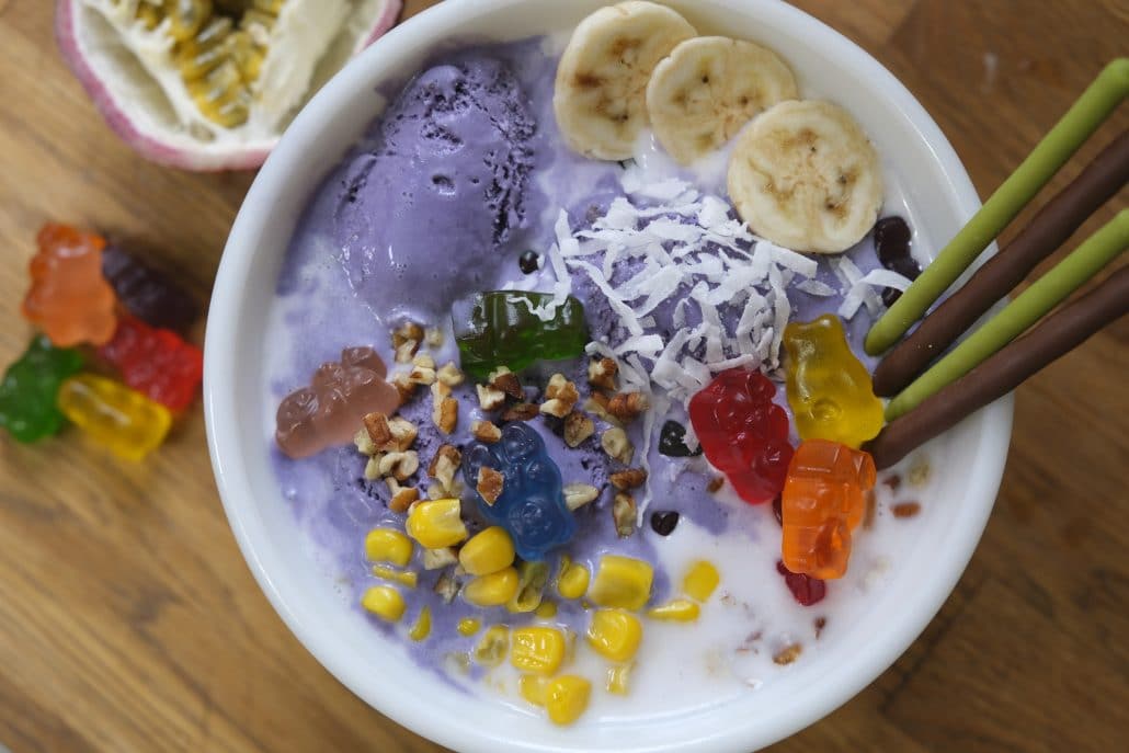 A bowl of halo-halo with pocky, banana slices, ube ice cream, gummy bears, and corn